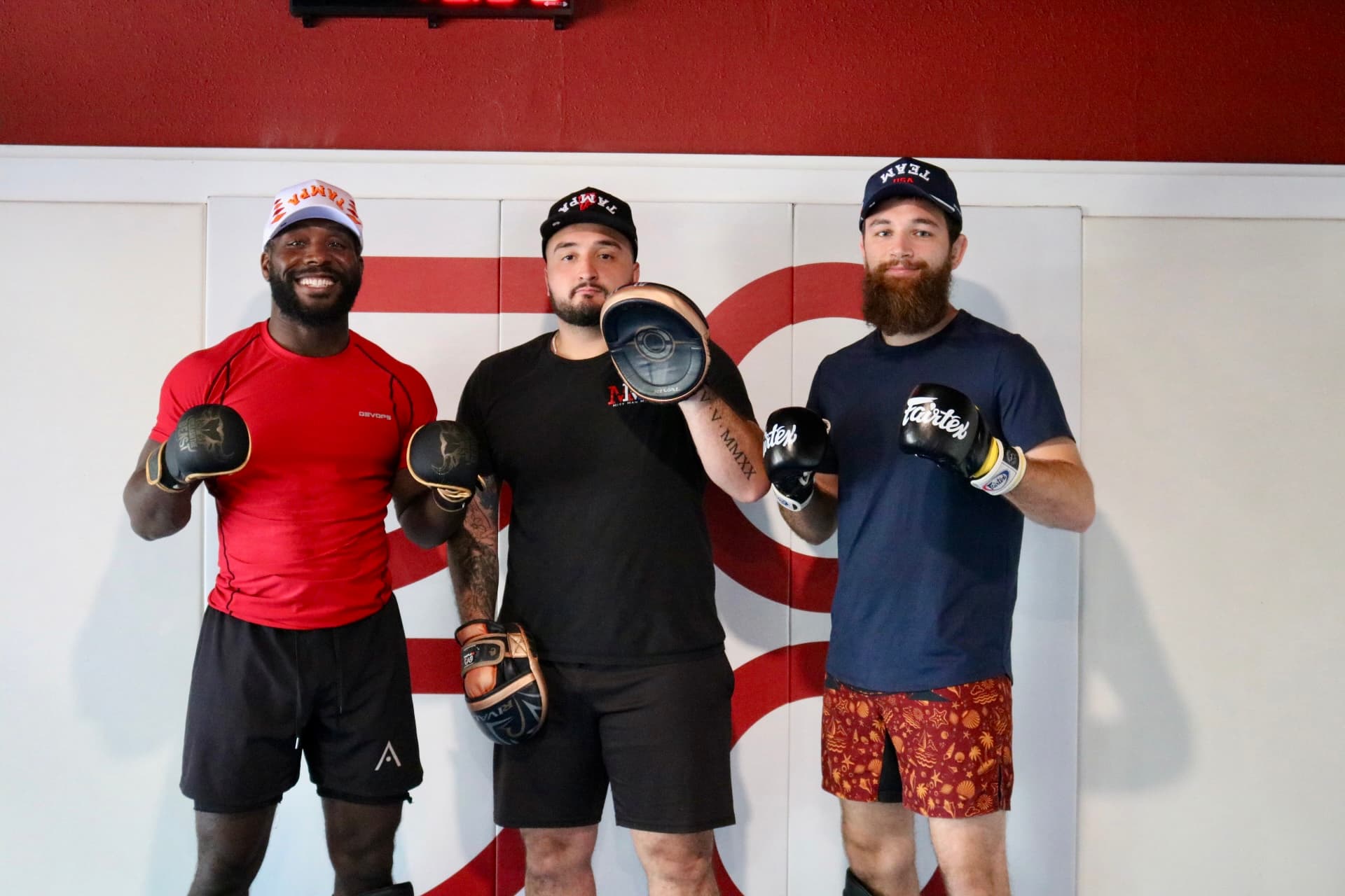 One striking coach with two of his fighters he is training holding up boxing mitts and gloves in front of a white wall.