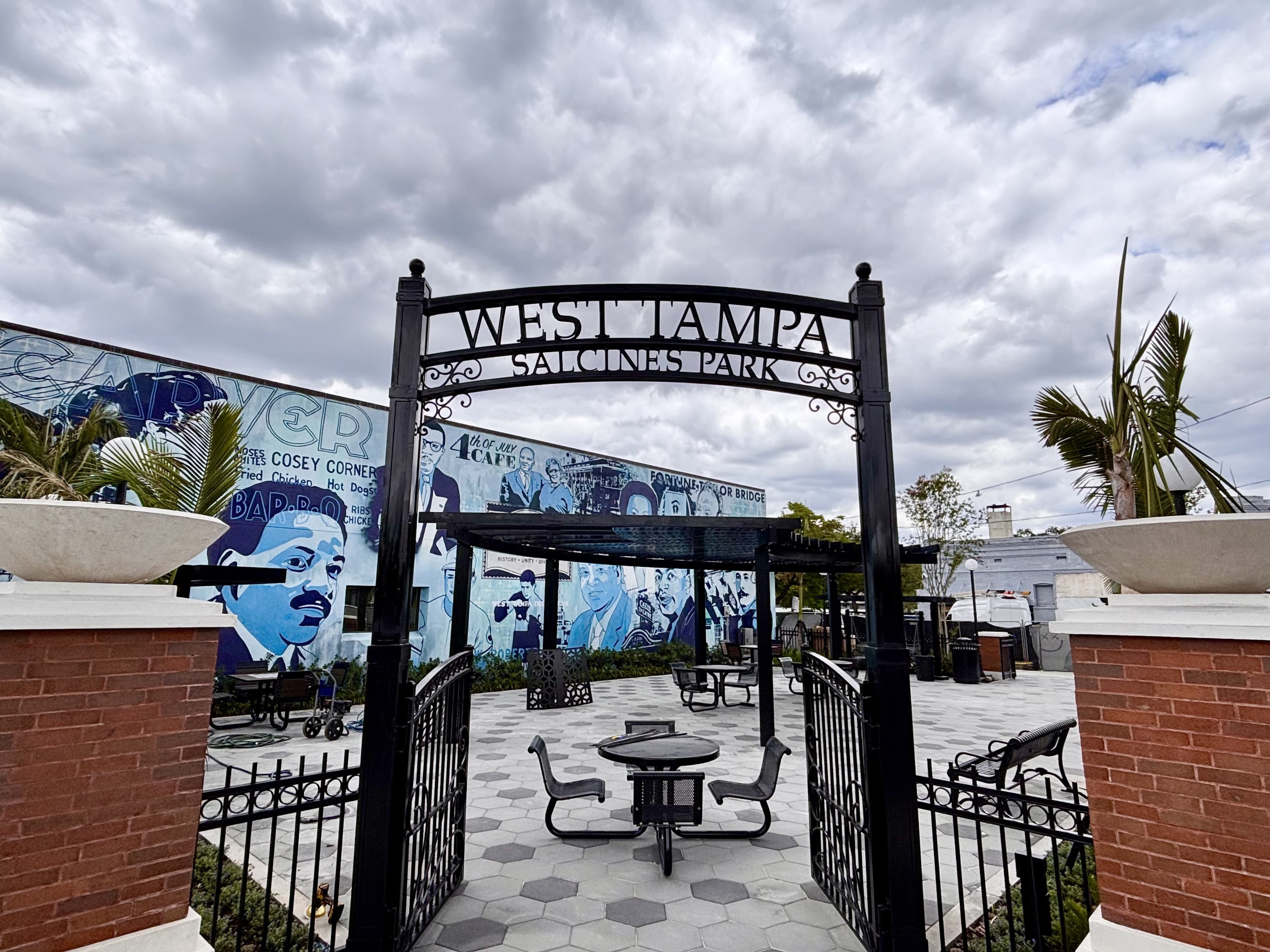 Black arch opening reading West Tampa Salcines Park. The park has black chairs and fencing to match the architecture in the area. The park is in front of the blue and white mural of historic members.