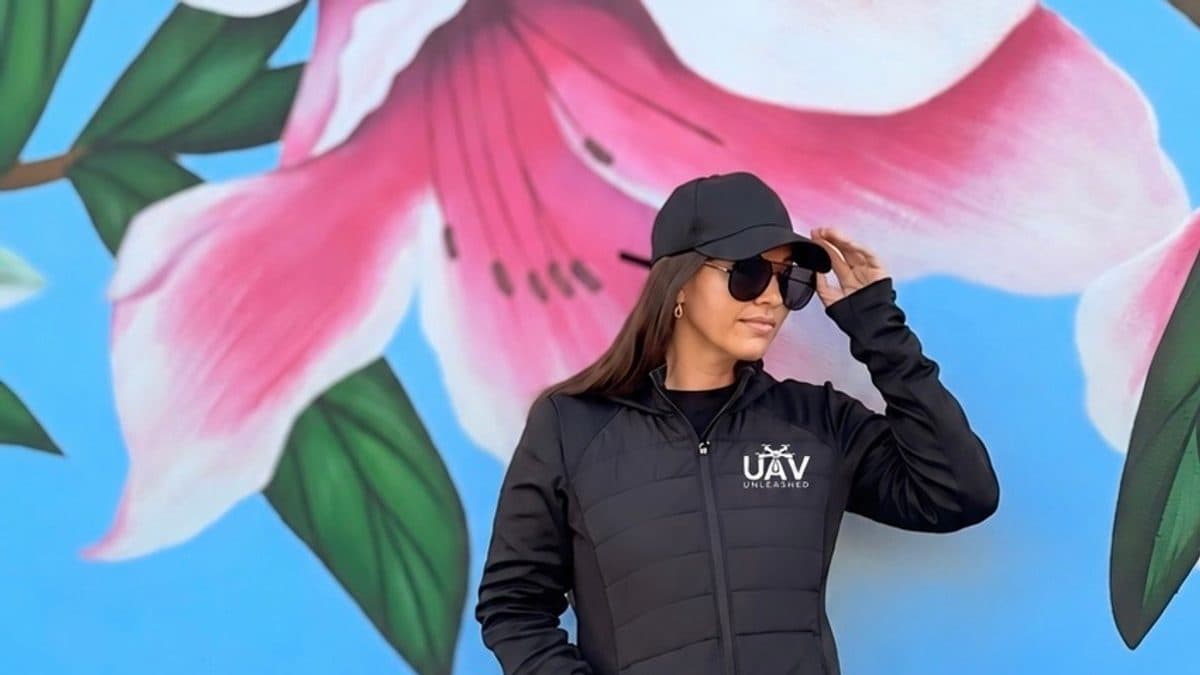 A woman wearing a black jacket and cap stands in front of a colorful pink flower mural, with the UAV Unleashed logo displayed