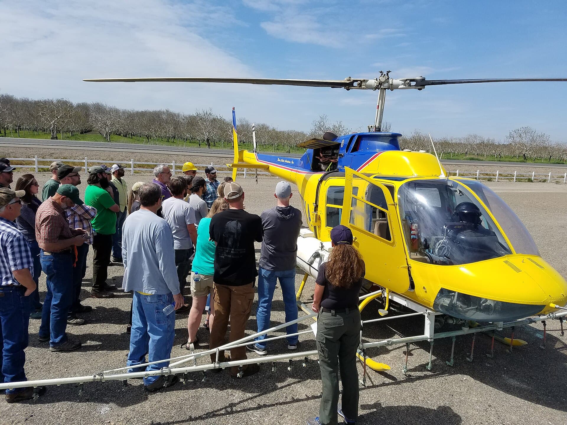 Yellow and blue spray helicopter with boom equipment parked on gravel during an aerial pesticide application training