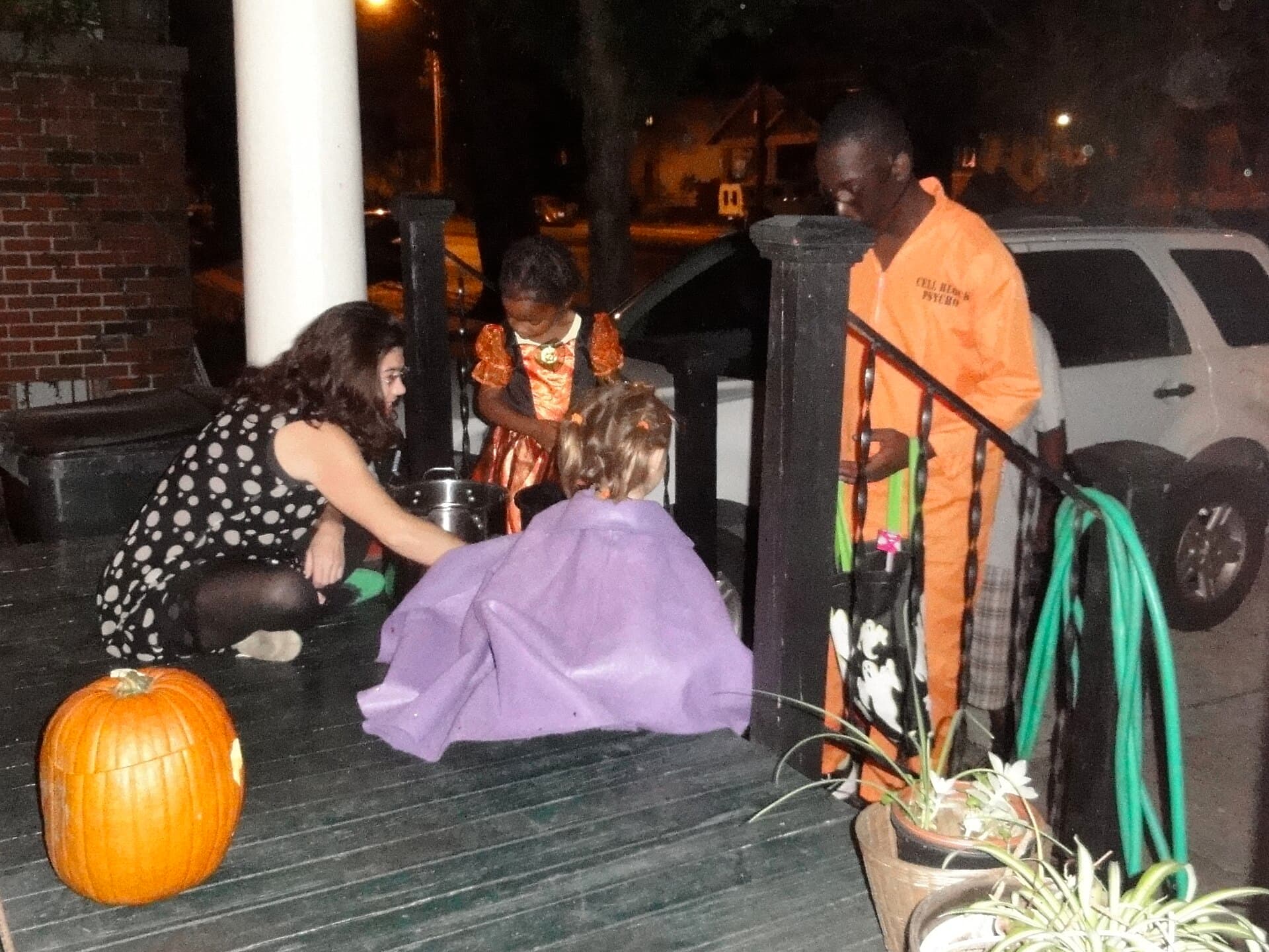 Children in Halloween costumes collecting candy on a front porch at night, with a pumpkin on the steps