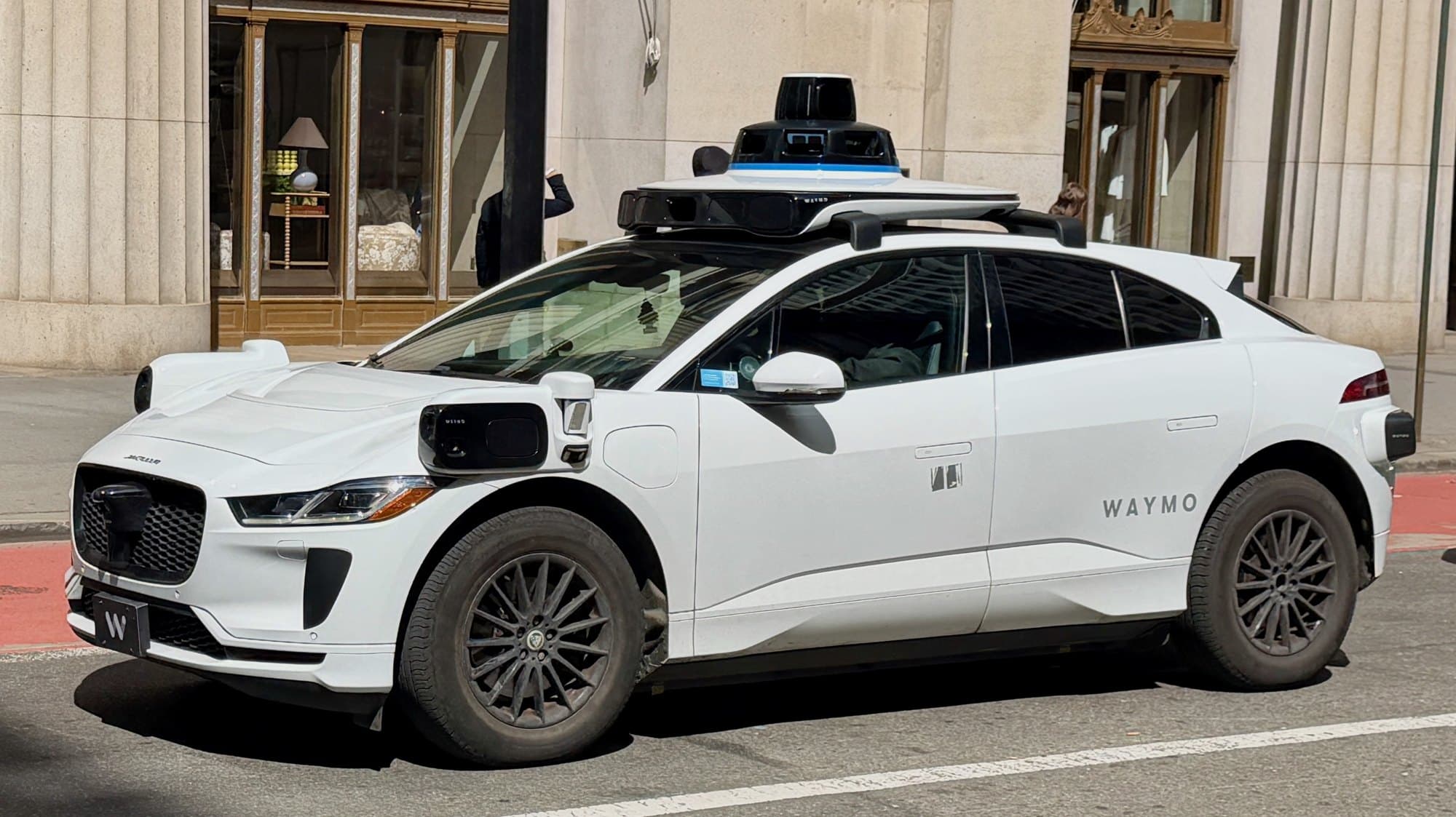 A white Waymo Jaguar I-PACE robotaxi with roof-mounted sensors parked at a curb